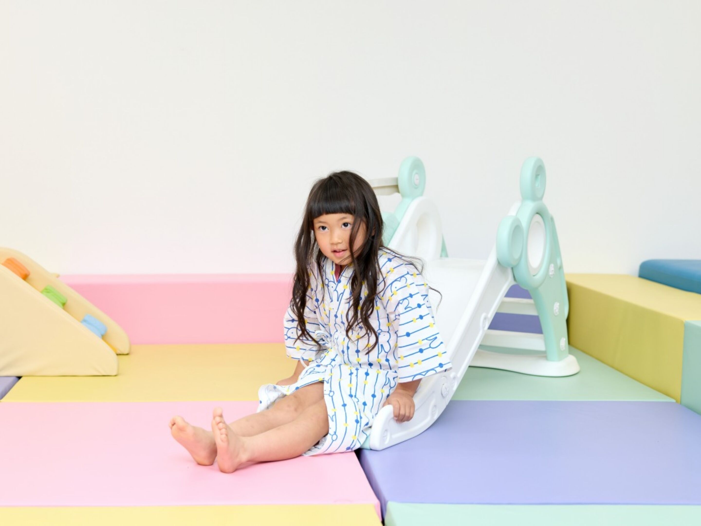 Un niño sentado sobre una alfombra de juego colorida en una habitación con pared blanca y una cama con colcha rosa y amarilla.