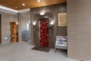 Un ascensor con puerta roja y pared de mármol. Se observa un cartel con los números de los pisos y una pequeña pantalla en la parte superior.