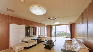 Amplia sala de estar con un sofá grande, una mesa de centro y un televisor. Cuenta con ventanas grandes con cortinas, un ventilador de techo y paneles de madera en las paredes.