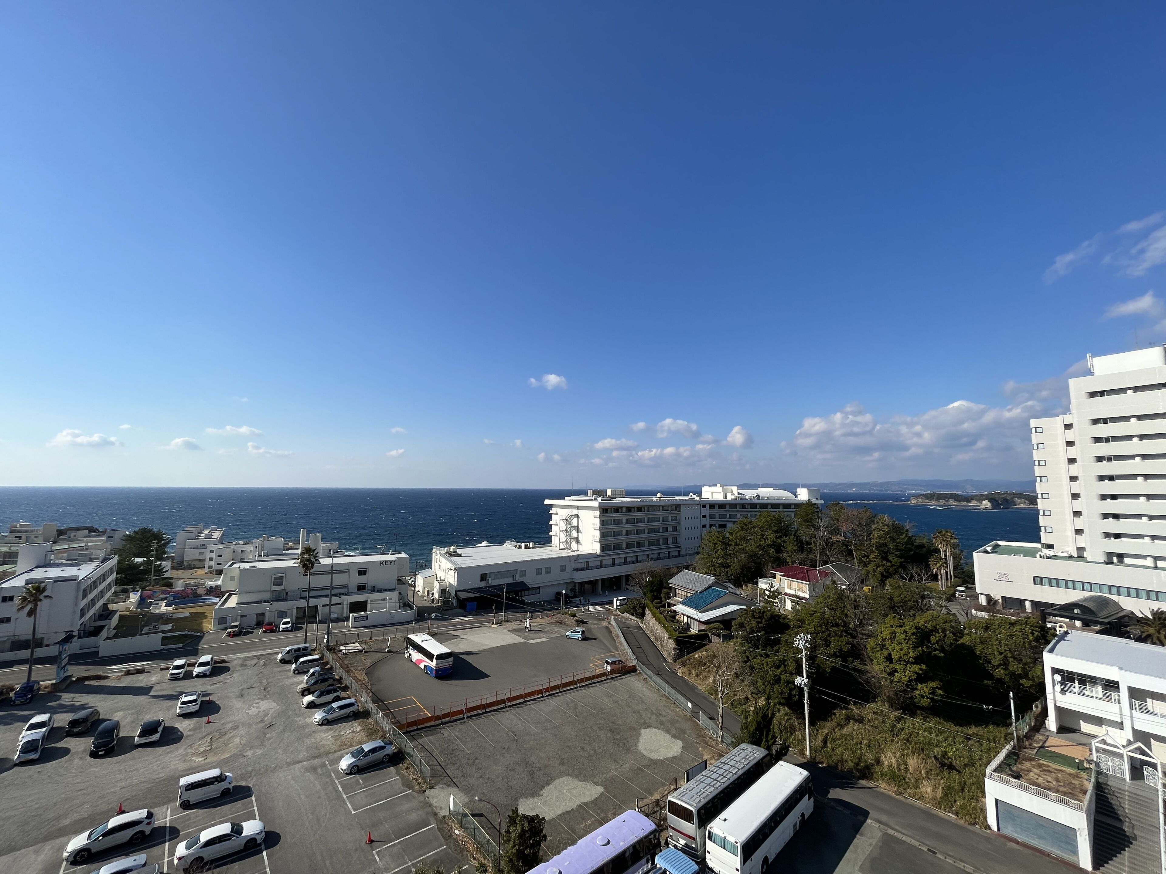 A parking lot with several cars and a bus, surrounded by buildings and a clear view of the sea.