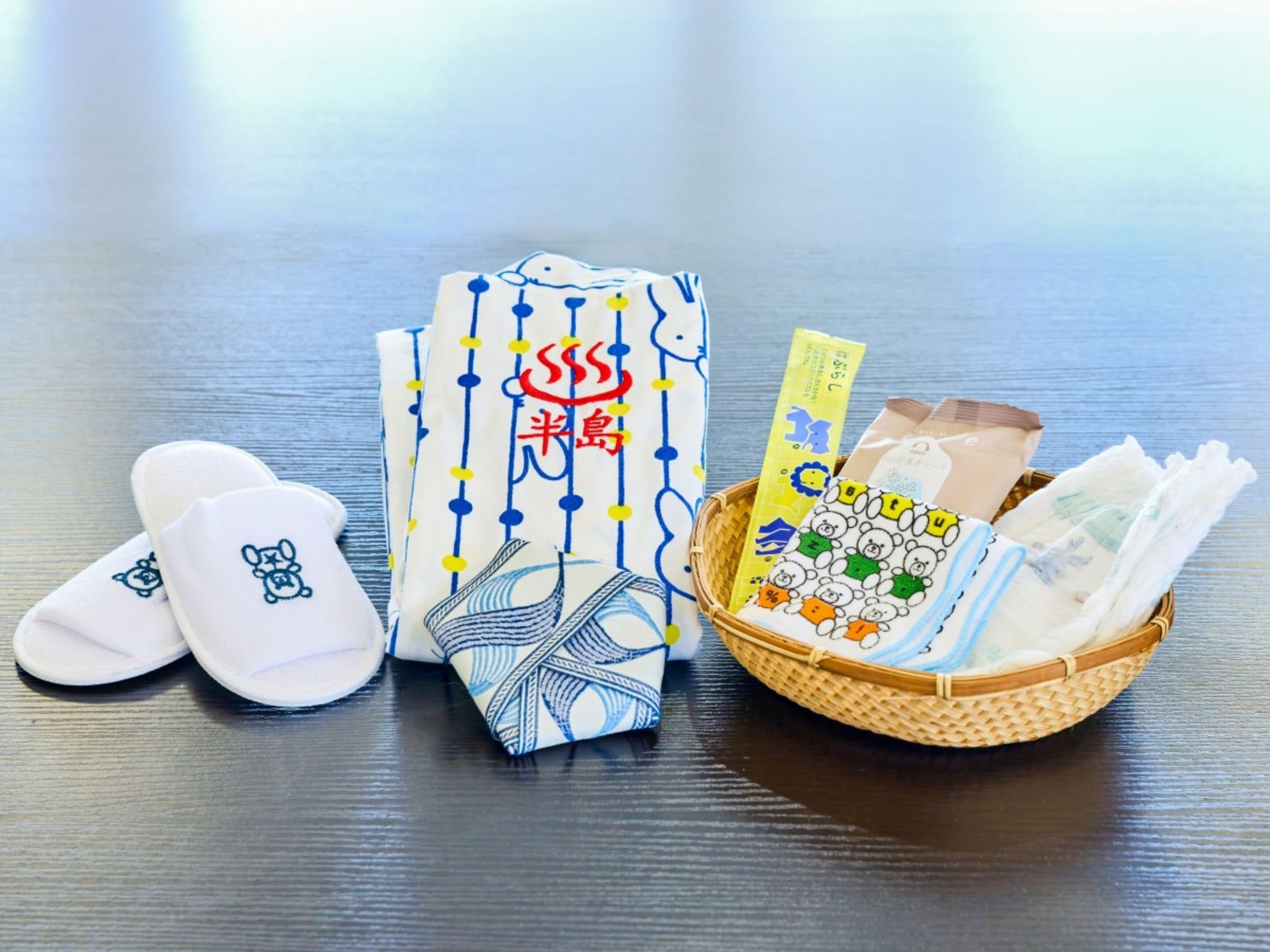 A table with a pair of white slippers, a patterned cloth, and a basket containing baby diapers and a card.