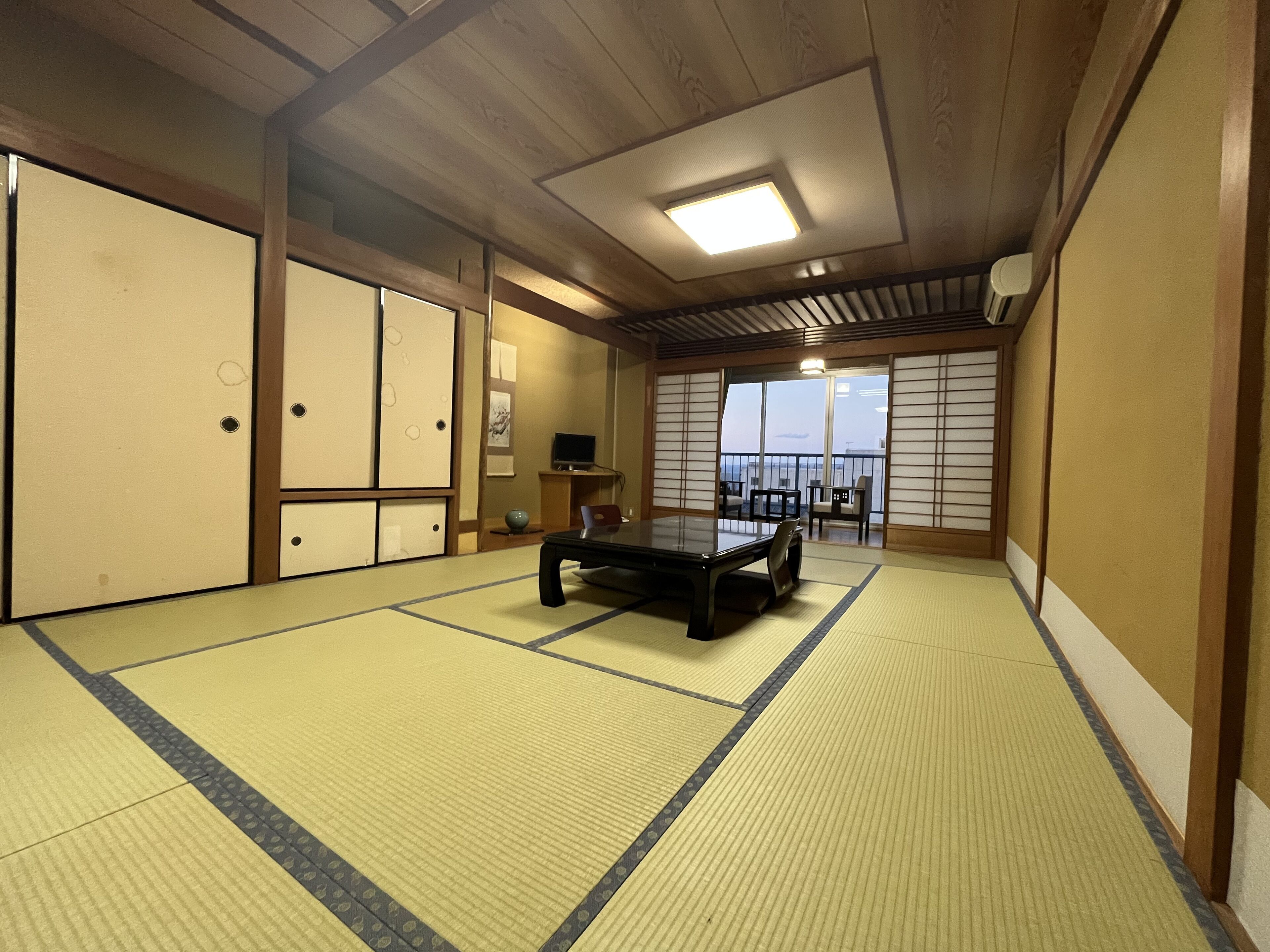 Une pièce de style japonais traditionnel, avec un sol recouvert de tatamis, une table basse, des portes coulissantes et une vue dégagée sur l’extérieur grâce à une grande fenêtre.
