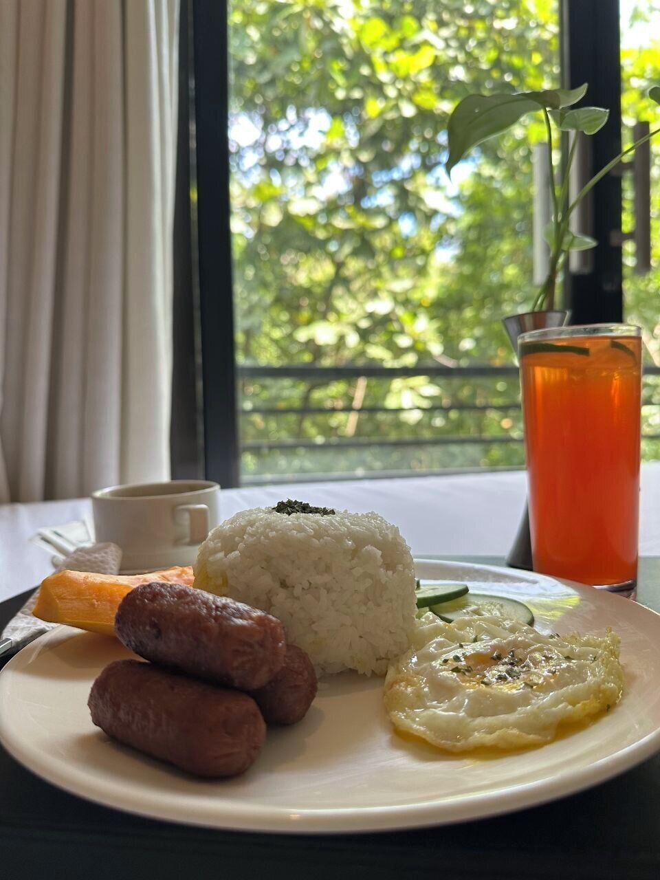 Um prato de arroz, salsichas e um prato com ovo, uma chávena e um copo de chá, com vista para o verde através de uma janela.