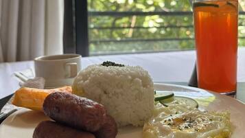 Um prato de arroz, salsichas e um prato com ovo, uma chávena e um copo de chá, com vista para o verde através de uma janela.