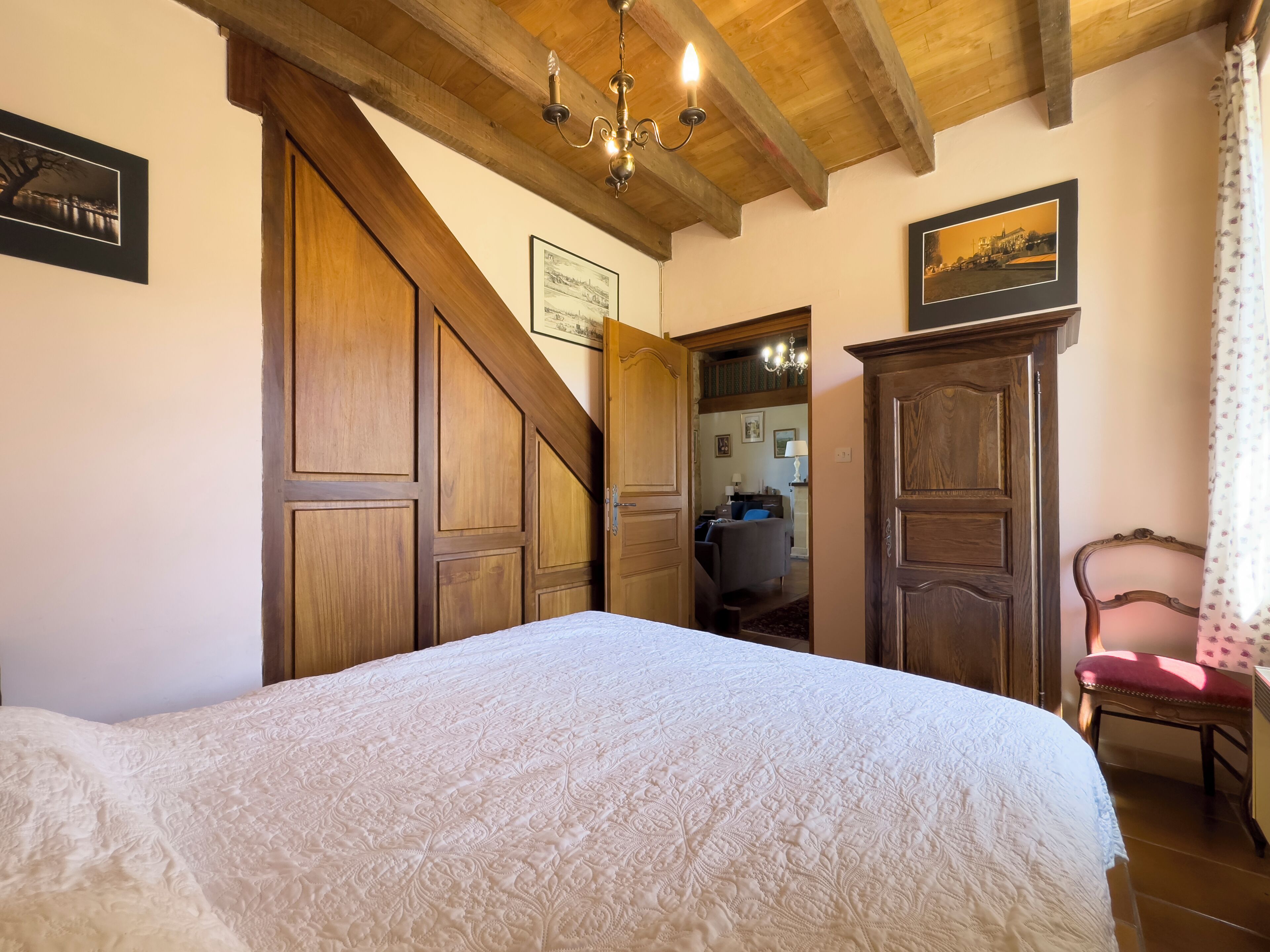Camera da letto con un letto grande, travi di legno a soffitto, un lampadario e vista su un'altra stanza con una scrivania e una sedia.