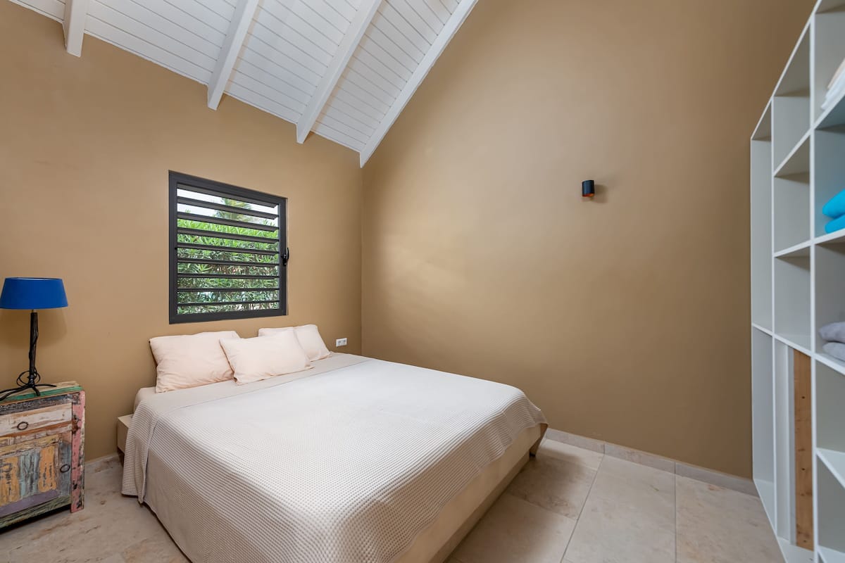 Une chambre à coucher comprenant un lit, une table de chevet, une lampe, une étagère et une fenêtre avec des stores.