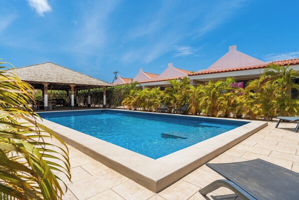 A swimming pool with a tiled deck, surrounded by palm trees and a covered area with a tiled roof.