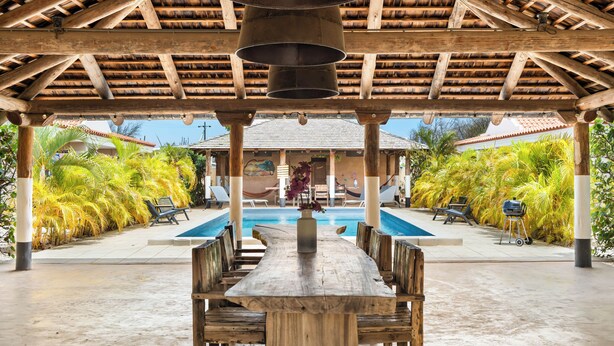 A wooden pergola with a pool table and chairs underneath, surrounded by palm trees and a view of a building with a tiled roof. - Kura Kabana by Boutique Bonaire Unique Resorts (Kralendijk)