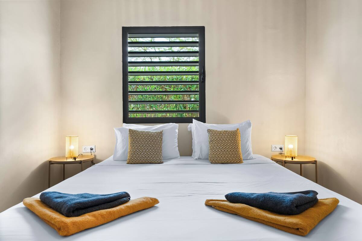 Une chambre d’hôtel avec un grand lit, deux tables de chevet avec des lampes, une fenêtre avec des stores et une vue sur la verdure.