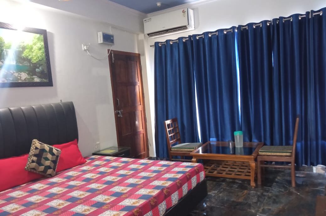 Un dormitorio con una cama, una silla de madera, una mesita pequeña, una cortina azul y un equipo de aire acondicionado.