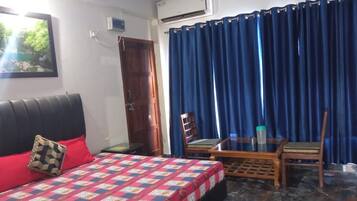 Un dormitorio con una cama, una silla de madera, una mesita pequeña, una cortina azul y un equipo de aire acondicionado.