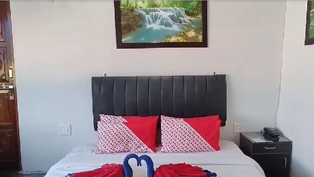 Un dormitorio con una cama, dos almohadas rojas con estampado, una mesita de noche con un teléfono y un cuadro colgado en la pared de una cascada.