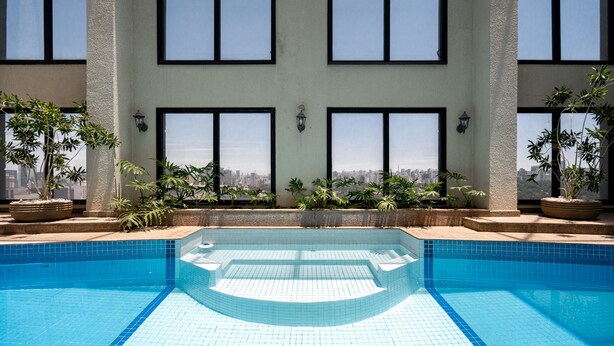 A swimming pool with clear blue tiles, surrounded by potted plants and large windows offering a city view. - Charlie Jerônimo da Veiga (São Paulo)
