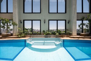 A swimming pool with clear blue tiles, surrounded by potted plants and large windows offering a city view.