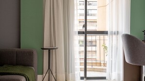 A room with a green wall, a grey sofa, a small table with a book, and a view of a balcony with a railing and a plant. - Charlie Jerônimo da Veiga (São Paulo)