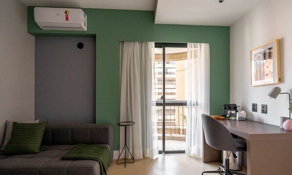 A modern room with a green wall, a grey sofa, a small table, a desk with a chair, and a window with curtains.