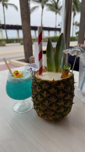 A pineapple cocktail with a straw and a small yellow duck float, and a glass of blue cocktail with a straw and a cherry on the rim, on a table with a view of palm trees and a building in the background.