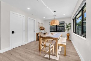 A modern dining area with a wooden table and chairs, a hanging light fixture, and a window with a view. - Coco Loco, tropical pool oasis near beach (Deerfield Beach)