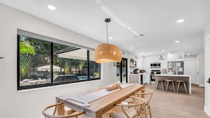 Eine moderne Küche mit einer großen Kochinsel, einem Essbereich mit Holztisch und -stühlen sowie einem großen Fenster mit Blick auf einen Garten.