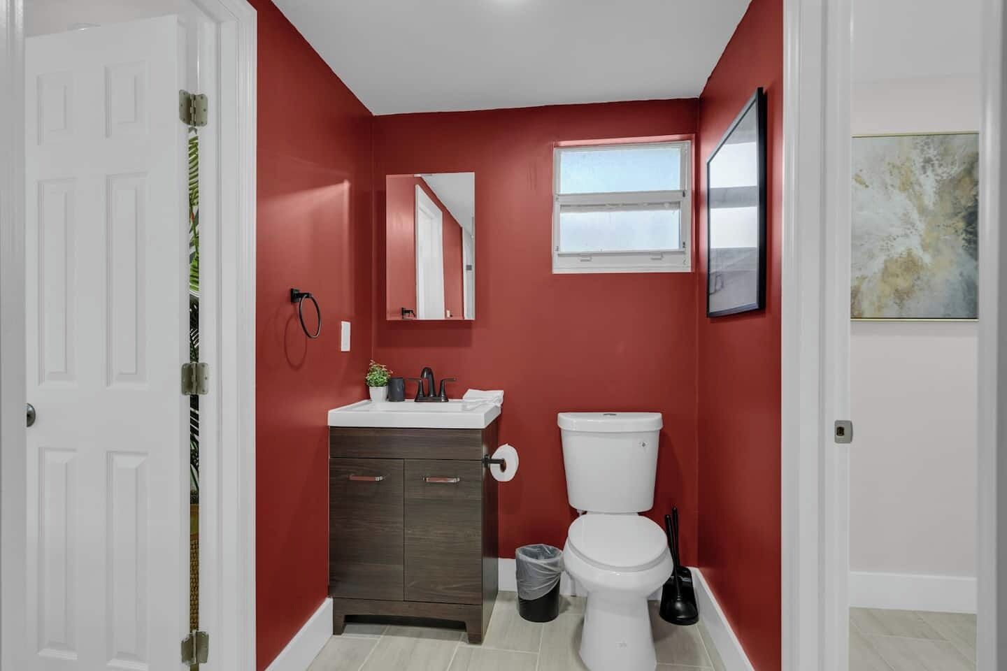 Une salle de bain avec des murs rouges, des toilettes blanches, un meuble-lavabo en bois foncé et une petite fenêtre.