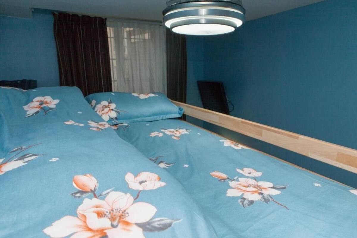 A bedroom with a floral bedspread, a wooden headboard, a ceiling light, and a window with curtains.