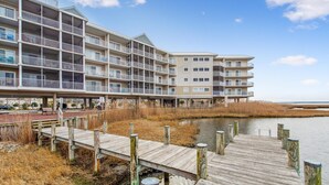 A multi-story hotel with balconies, a wooden dock extending over water, and a beach in the background.