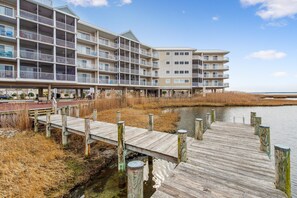 A multi-story hotel with balconies, a wooden dock extending over water, and a beach in the background.