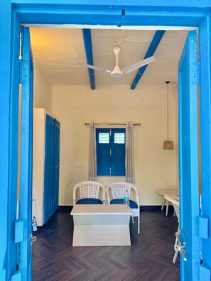 A room with a blue door, a ceiling fan, a window with curtains, two white chairs, a small table, and a woven basket. - Mykonos Villa (Varanasi)
