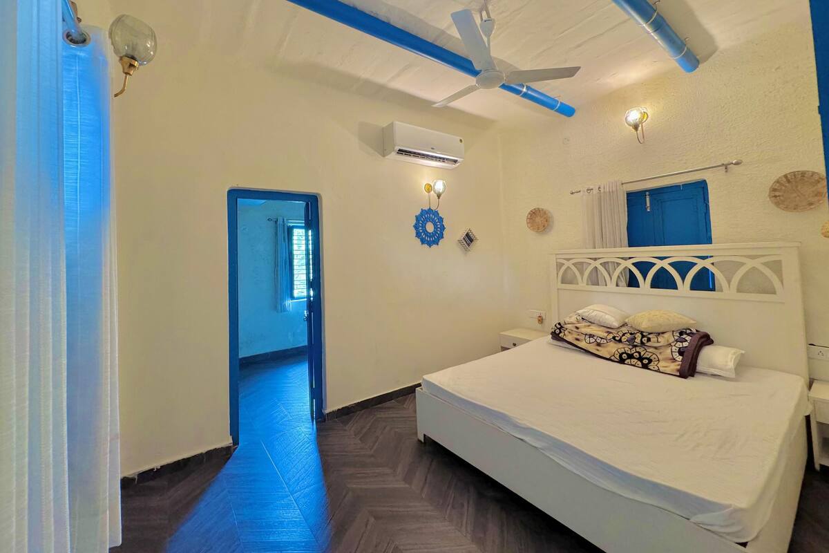 A bedroom with a bed, a ceiling fan, an air conditioning unit, a blue door, and a herringbone-patterned floor.