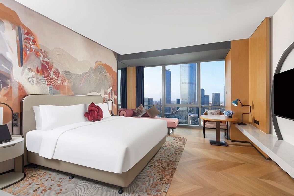 A modern hotel room with a large bed, a desk, and a view of the cityscape through large windows.