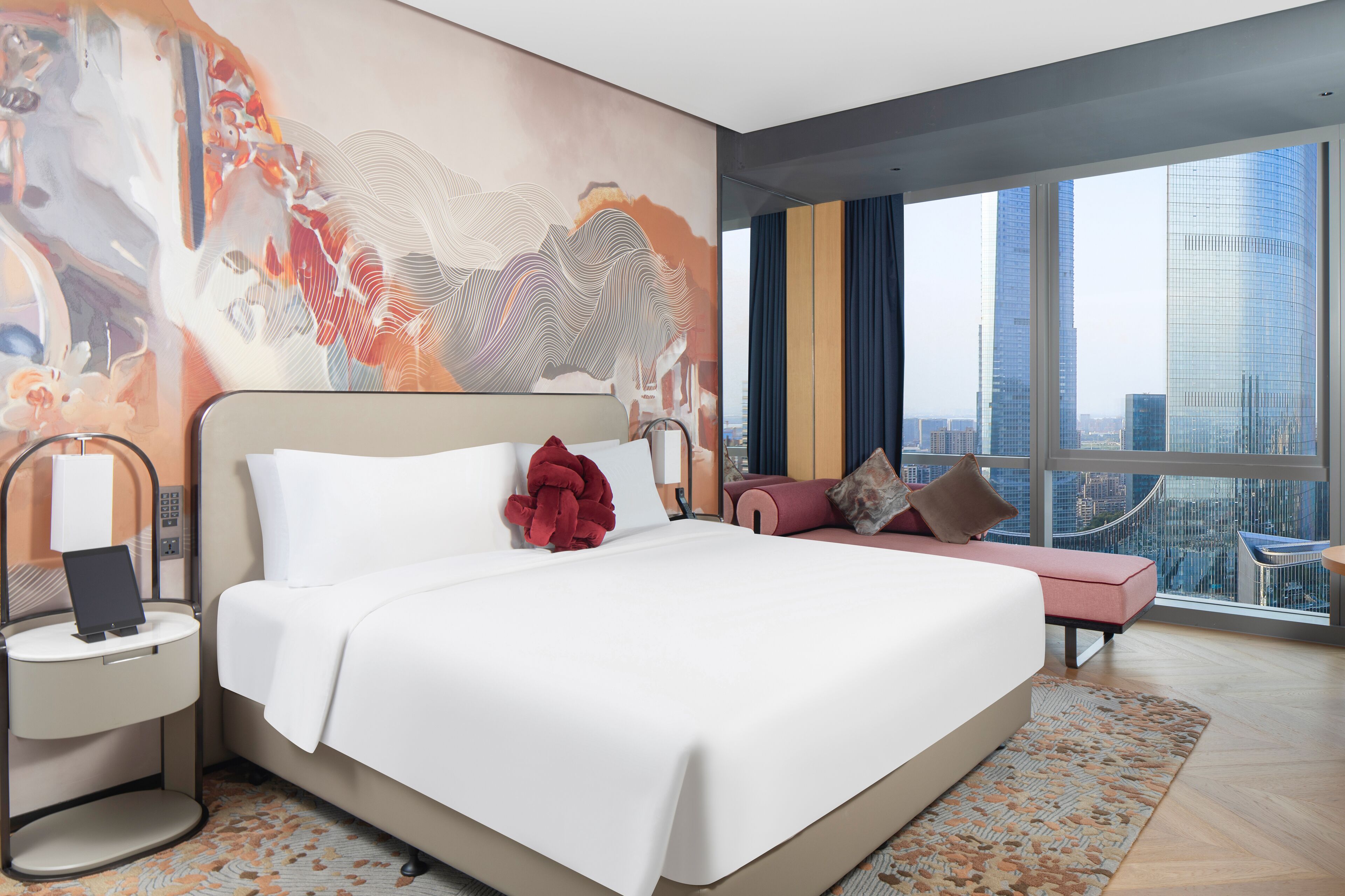Habitación de hotel moderna con una cama grande, una mesita de noche, un espacio para sentarse y vistas a la ciudad a través de amplios ventanales.