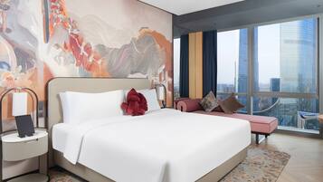 Habitación de hotel moderna con una cama grande, una mesita de noche, un espacio para sentarse y vistas a la ciudad a través de amplios ventanales.
