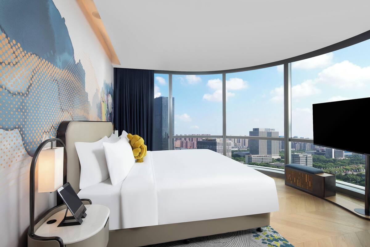 A modern hotel room with a large bed, a bedside table with a lamp, a TV, and a city view through large windows.