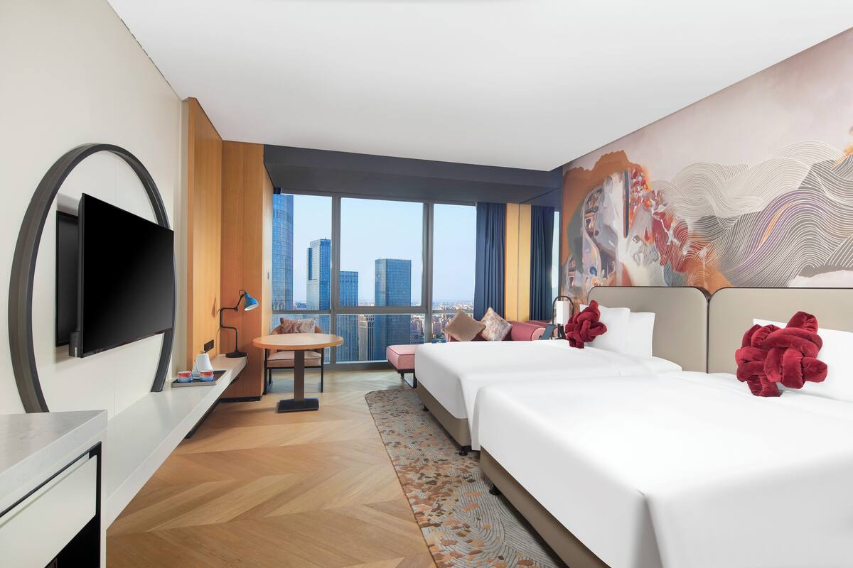 A hotel room with a large bed, a desk, a chair, a TV, and a view of the cityscape.
