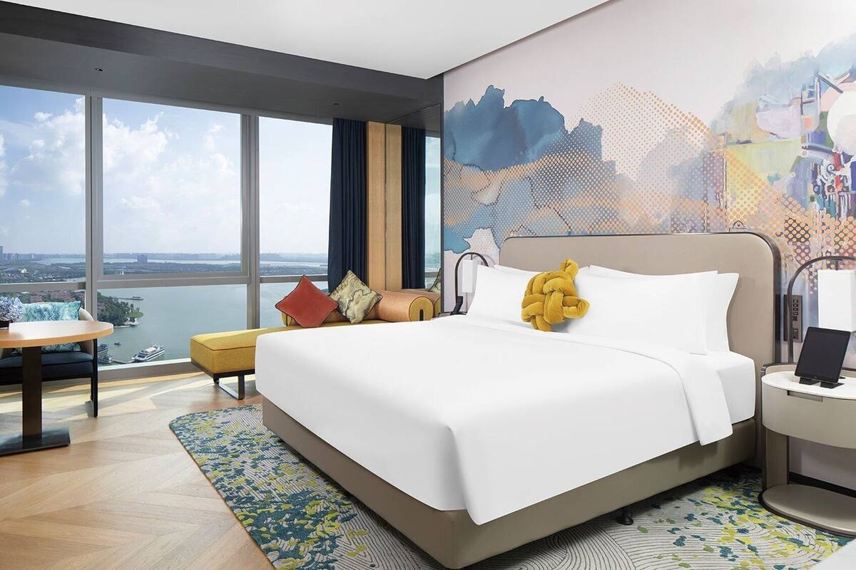 A modern hotel room with a large bed, a desk, and a view of a cityscape through large windows.