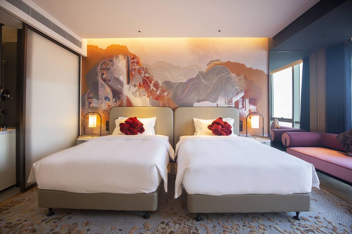 A hotel room with two beds, a large wall mural, a sofa, and a window.