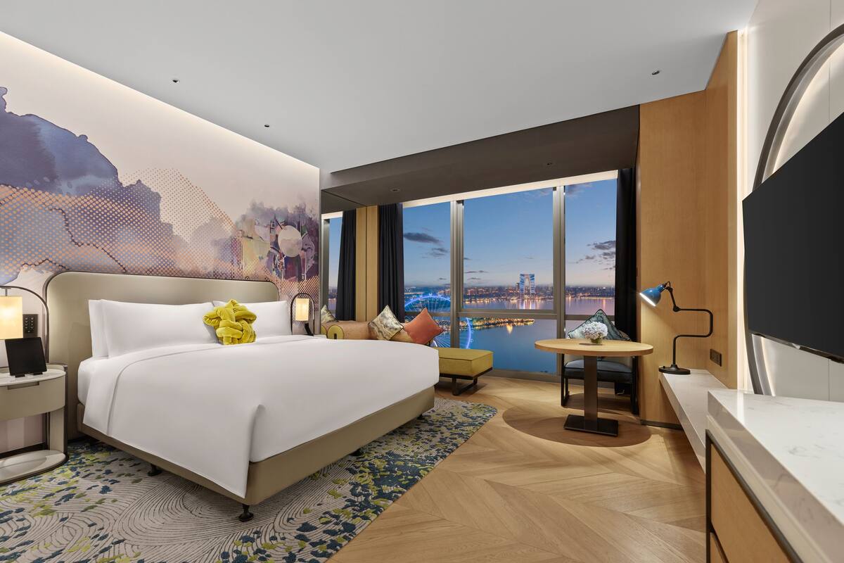 A modern hotel room with a large bed, a desk, a small table, and a view of a cityscape through large windows.