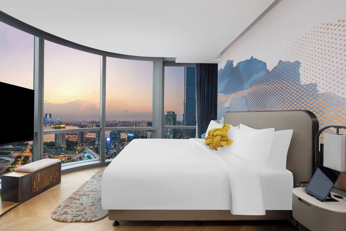 A modern hotel room with a large bed, a desk with a laptop, and a view of a cityscape at sunset.
