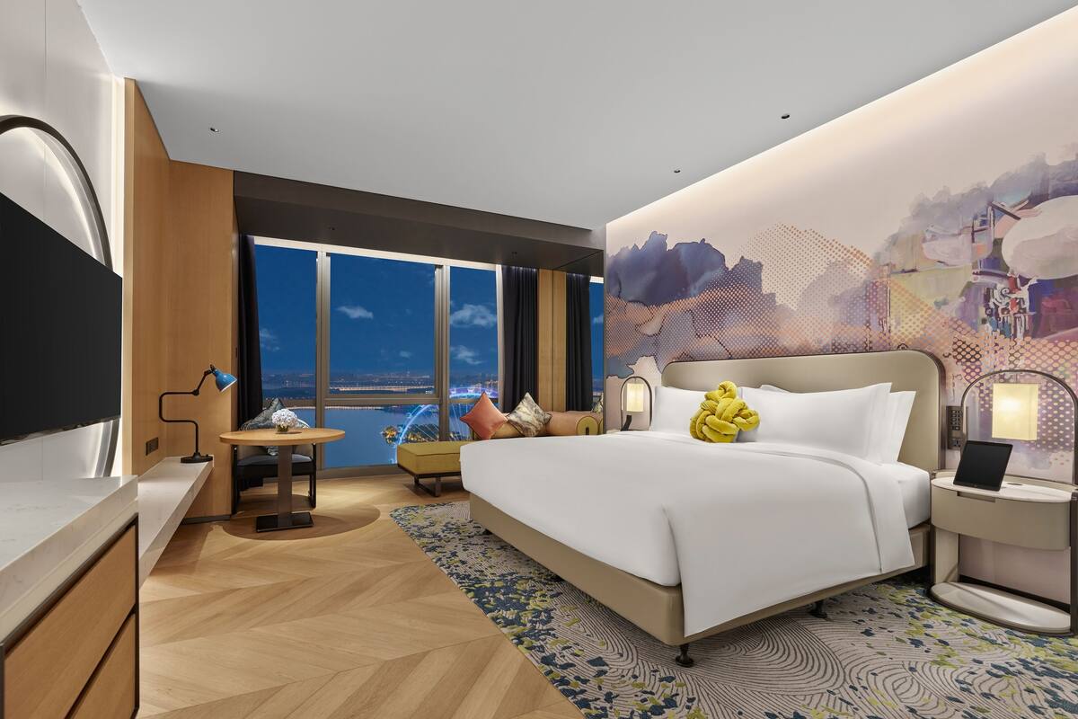 A modern hotel room with a large bed, a desk, a chair, and a view of the cityscape through large windows.