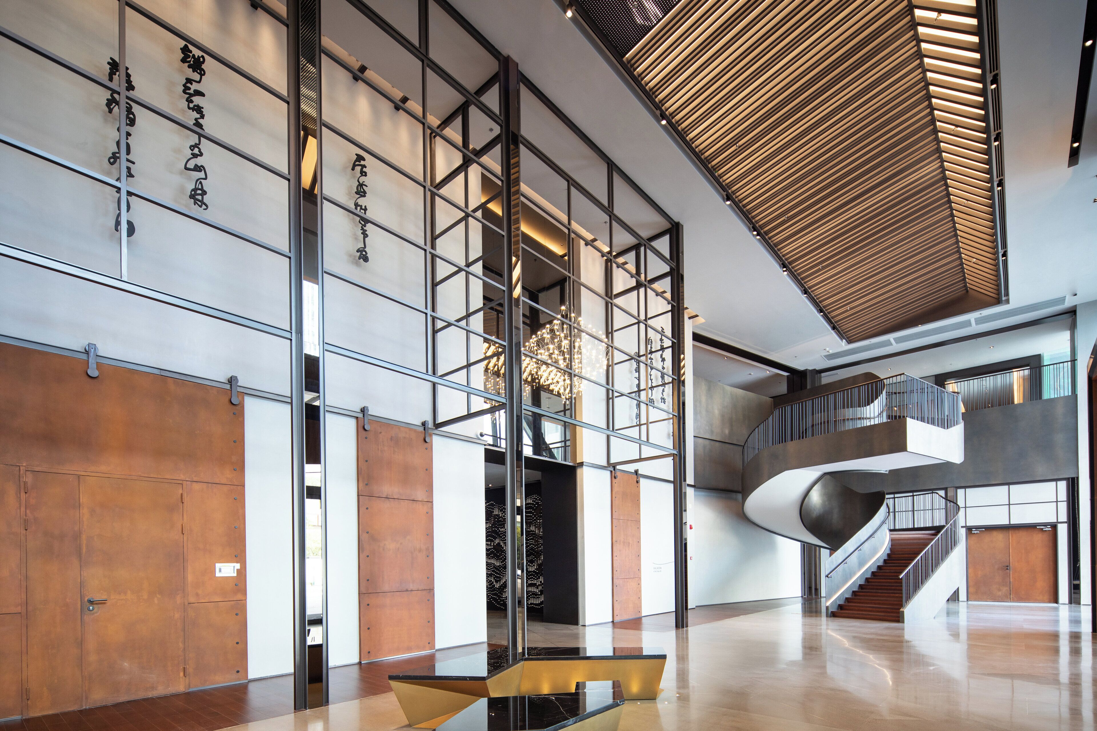 A spacious lobby with a modern design, featuring a central staircase, a wooden slatted ceiling, and a large chandelier.