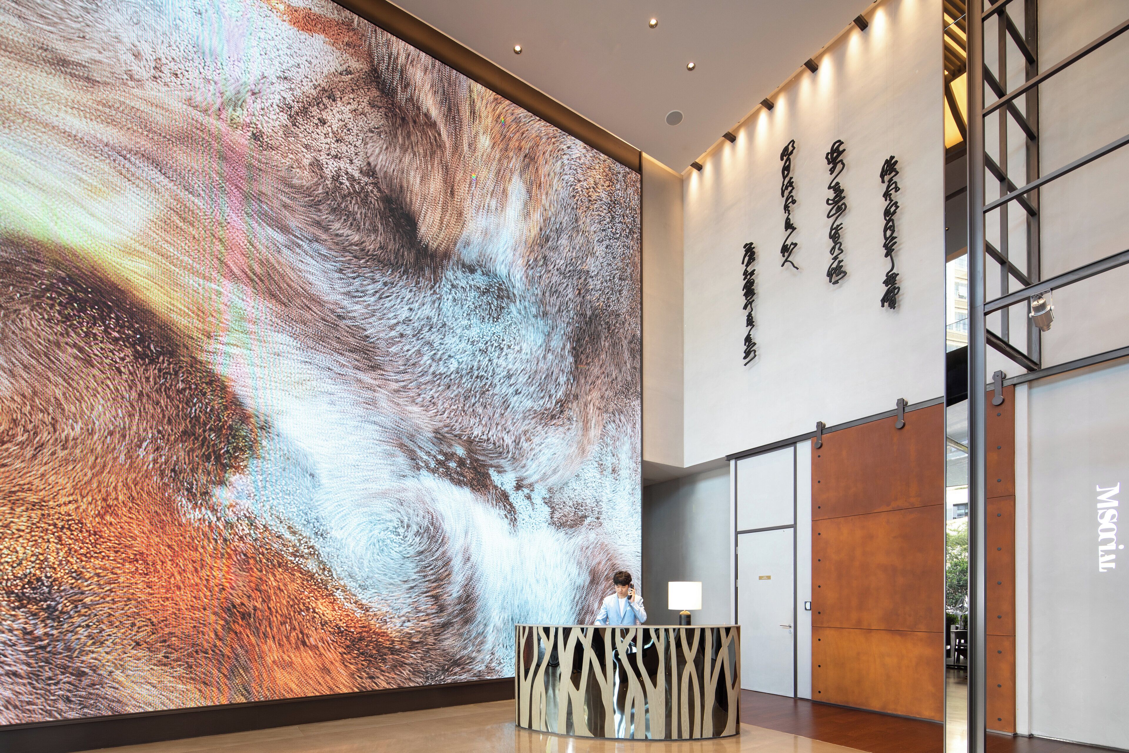 A modern hotel lobby with a large digital display, a reception desk, and a wall with text.