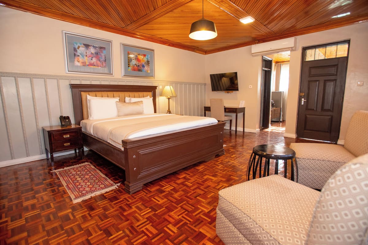 A hotel room with a wooden floor, a bed with white linens, a desk with a chair, a TV mounted on the wall, and a door leading to another room.