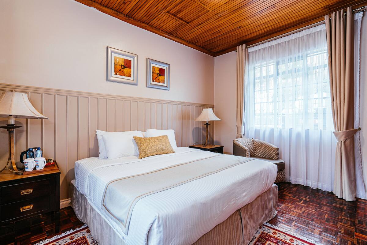 A hotel room with a large bed, two bedside tables, a chair, a window with curtains, and two framed pictures on the wall.