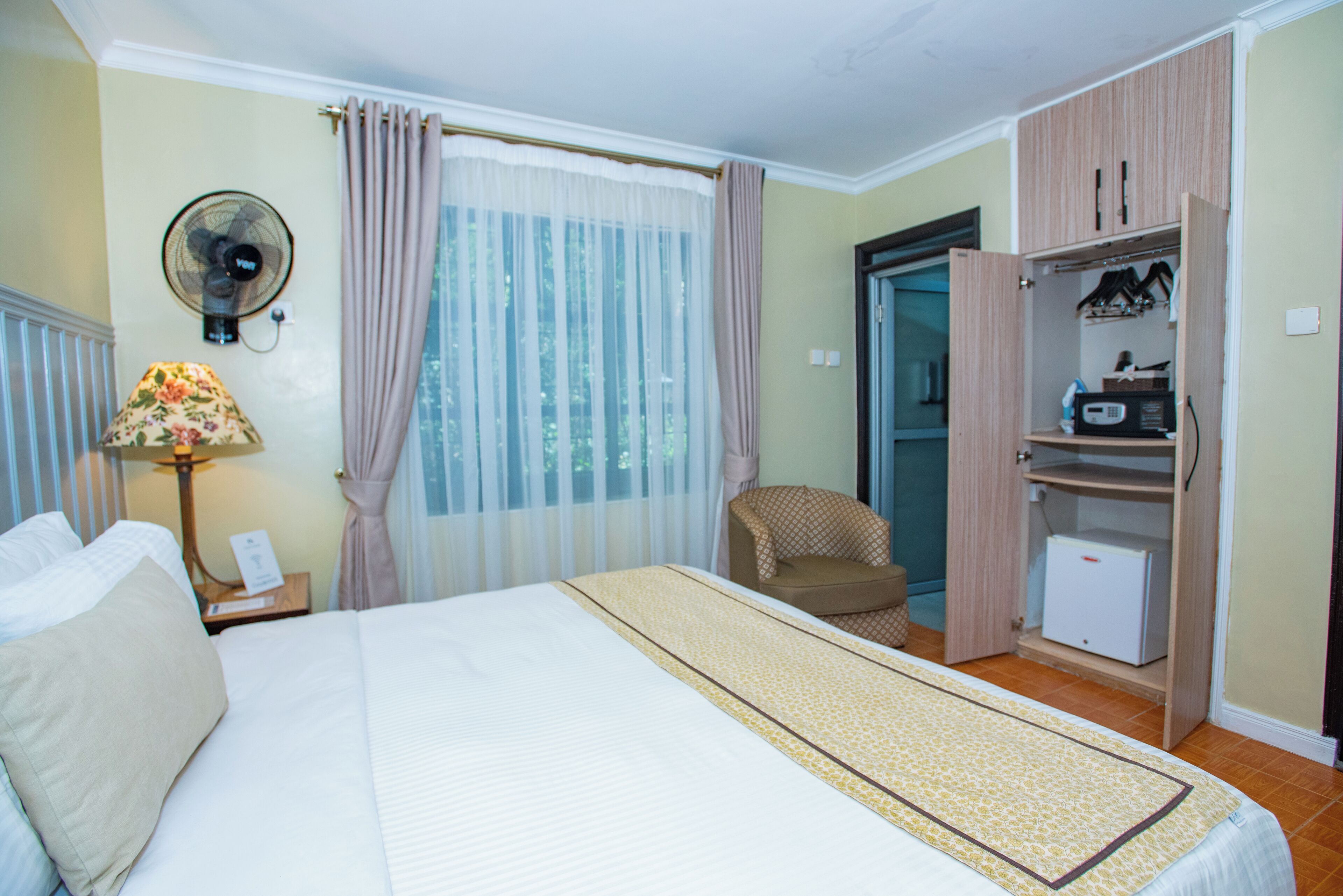 Habitación de hotel con cama, escritorio con lámpara, silla, armario y un mueble pequeño.