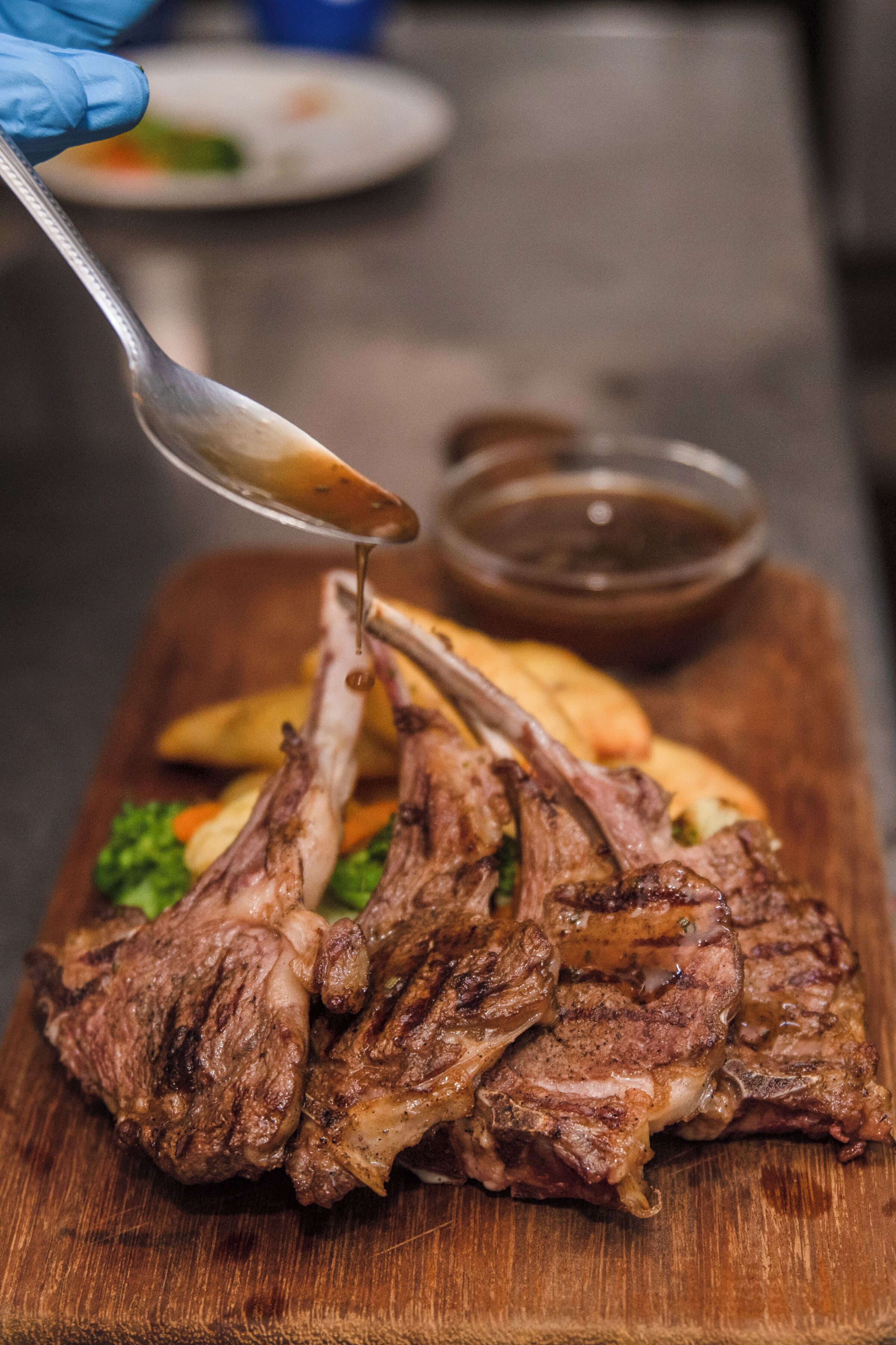 A wooden cutting board with grilled lamb chops and a spoonful of sauce being poured over them.