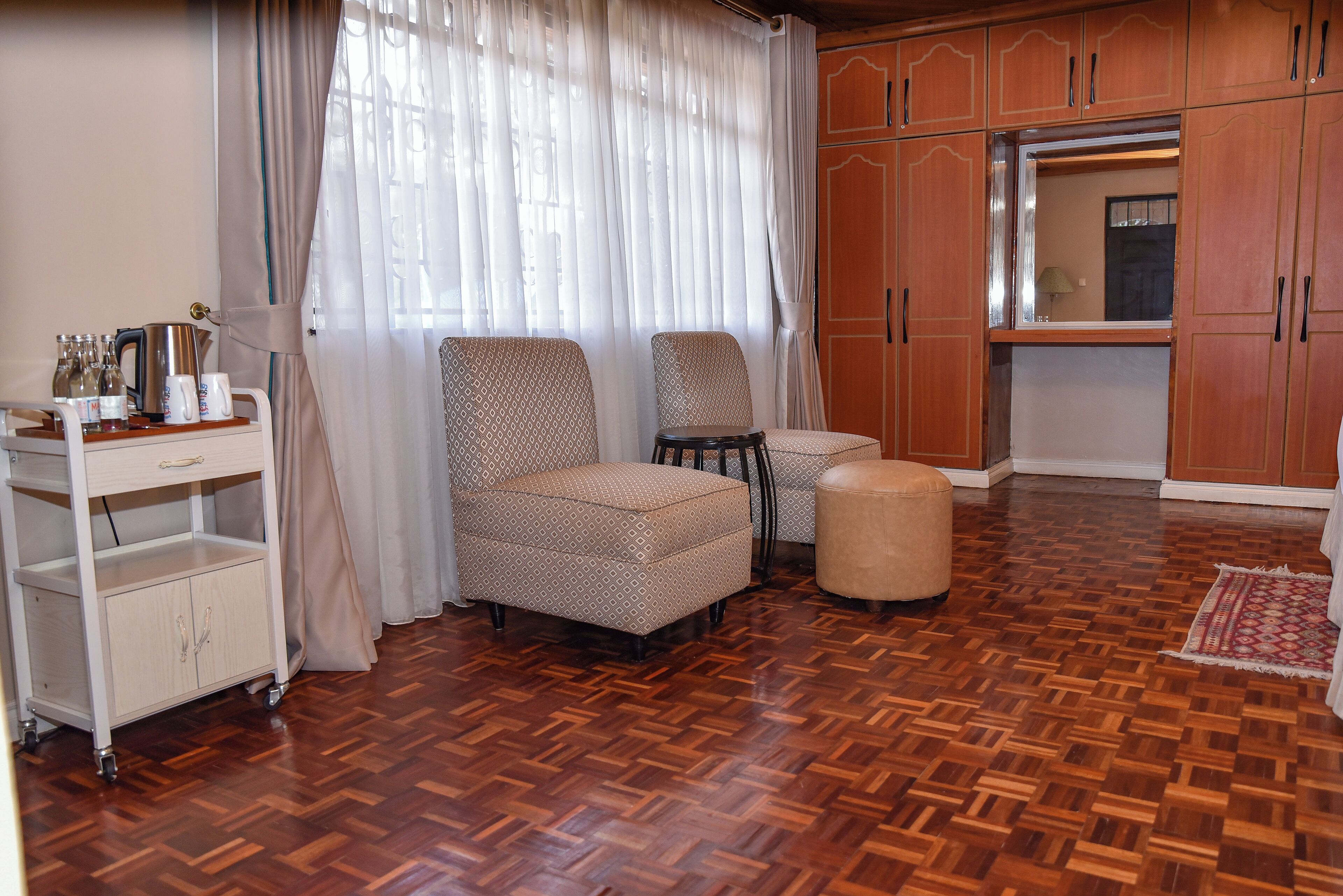 Ein Zimmer mit Holzdielenboden, zwei gemusterte Sessel, ein kleiner Hocker, eine weiße Kommode mit Schubladen und ein Servowagen mit Flaschen und einem Wasserkocher.