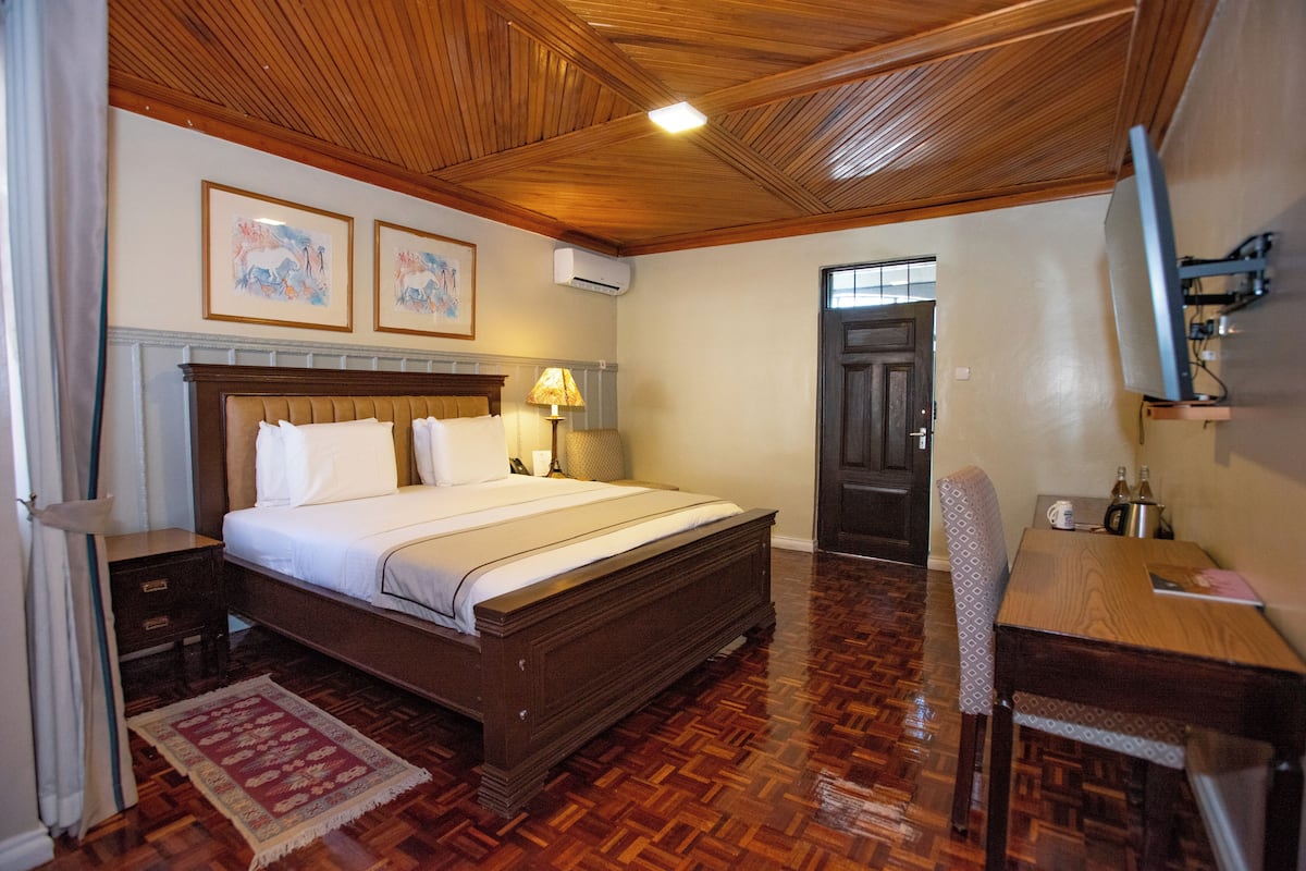 A bedroom with a wooden bed, bedside tables, a desk, and a chair. There are two framed pictures on the wall, an air conditioning unit, and a door leading to another room.