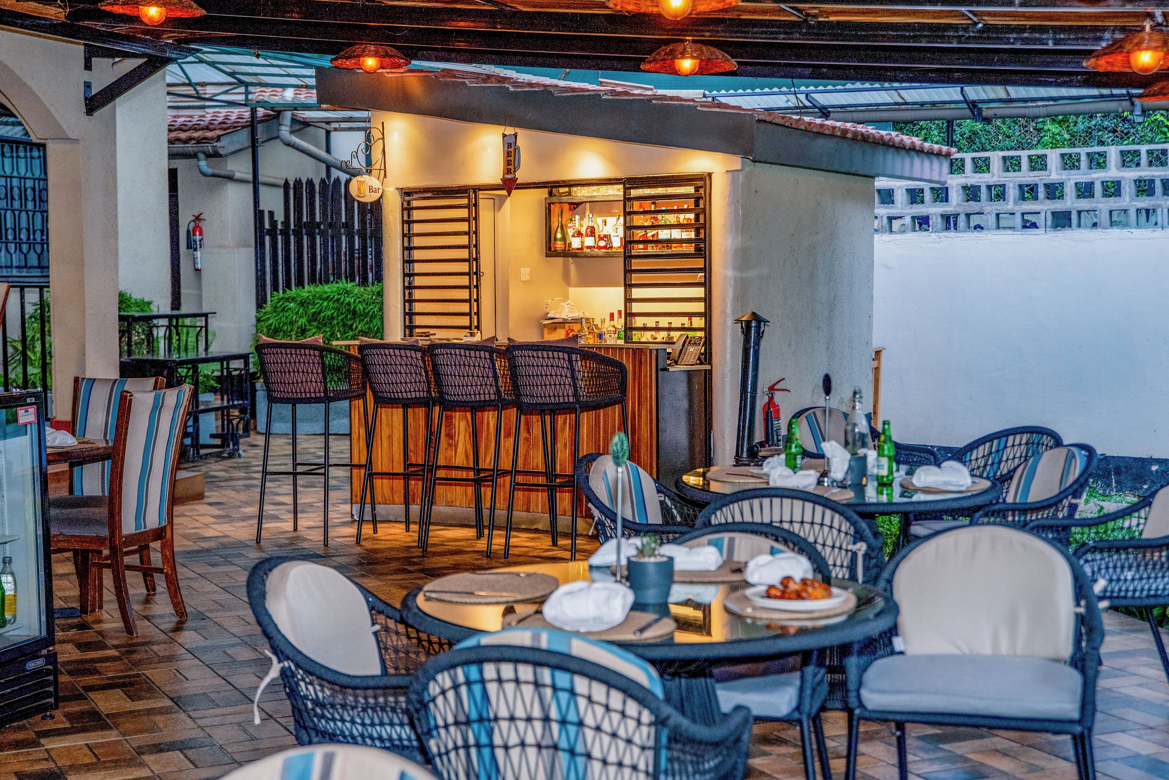 An outdoor dining area with wicker chairs and tables, a bar area with stools, and a view of the surrounding greenery.
