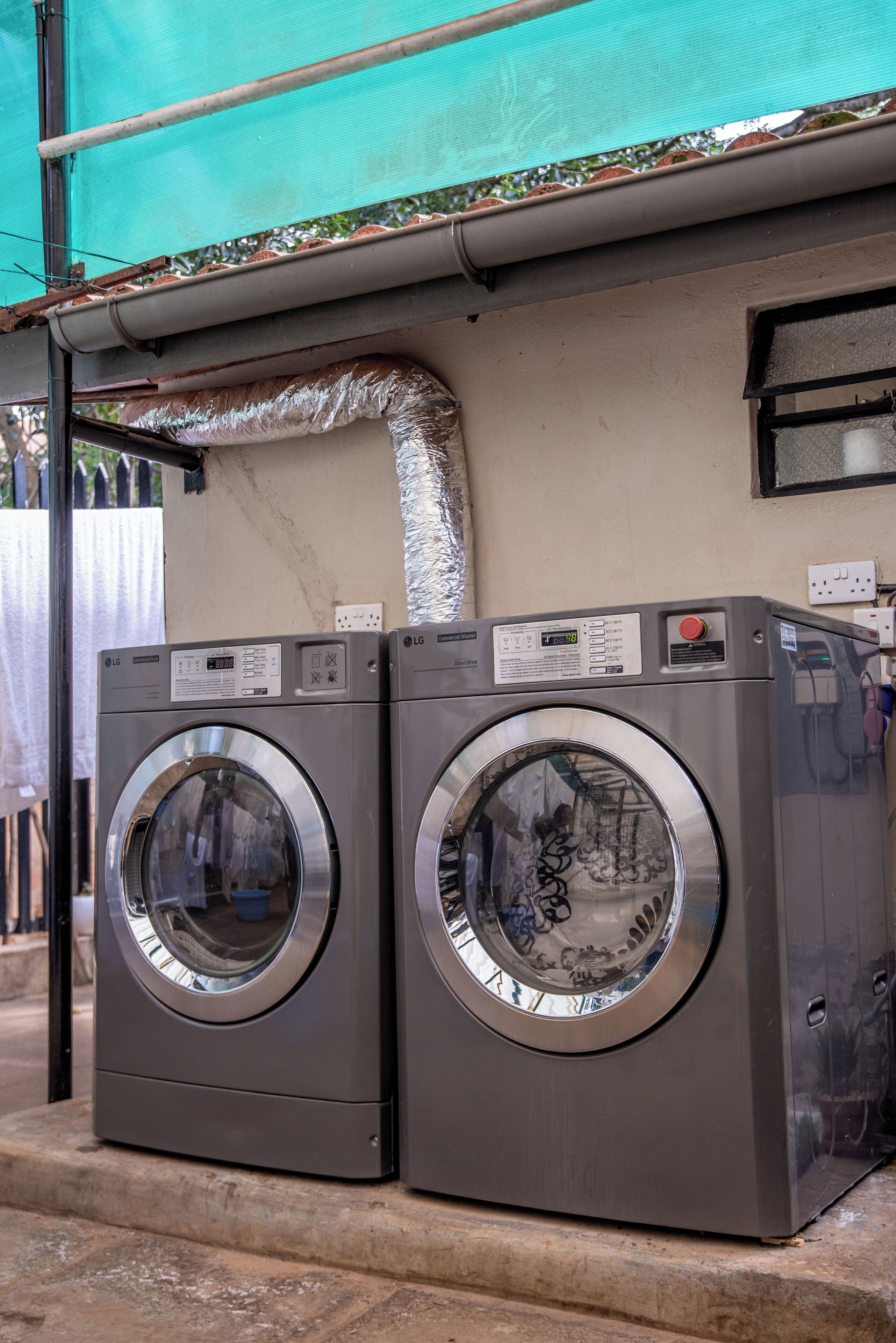 Two front-loading LG washing machines installed outdoors, with a green canopy overhead and a metal exhaust pipe above them.