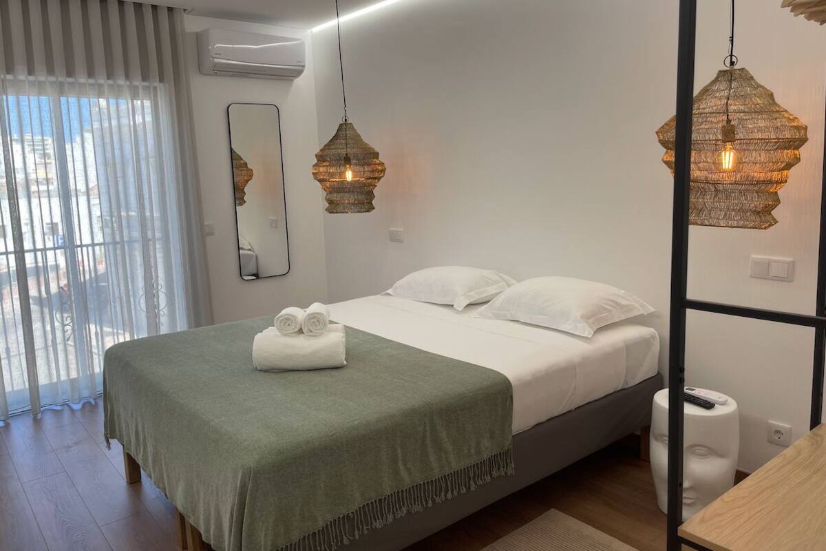A modern bedroom with a bed, two hanging lamps, a mirror, and a window with blinds.